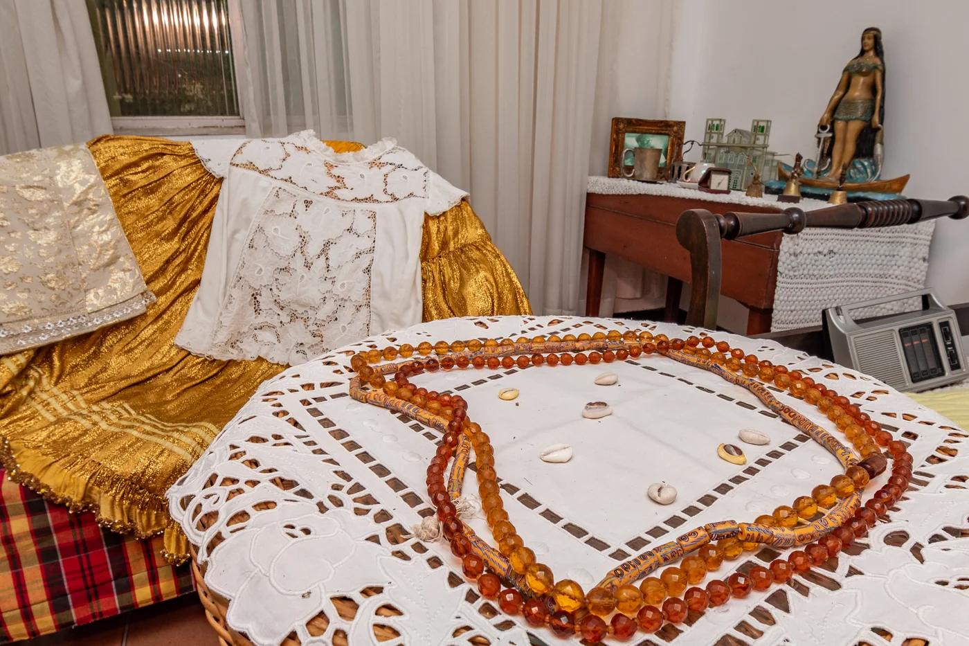 Joias de axé com mesa do oráculo, vestimentas com saia, bata, pano da costa e ojá, e outros elementos do cotidiano de Mãe Menininha;