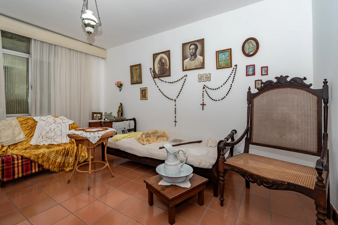 Visualização do trono, cama, mesa do oráculo, peça com objetos e imaginária, bengala, xale, rádio, valise com utensílios pessoais, fotografias e crucifixos.
